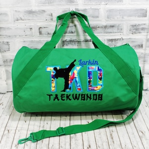 May include: Green duffel bag with a black silhouette of a person doing a taekwondo kick. The bag has the text "TKD" in colorful letters with the word "Taekwondo" underneath. The name "Larkin" is written above the "TKD" text.