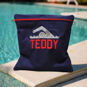 May include: Navy blue tote bag with a red zipper and the name "TEDDY" embroidered in red. The bag features a silver swimming graphic above the name. The bag is sitting on a stone ledge next to a swimming pool.