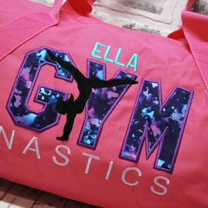 May include: Pink gym bag with a purple and blue "GYM" design with a silhouette of a gymnast in the middle. The text "ELLA GYMNASTICS" is embroidered on the bag.