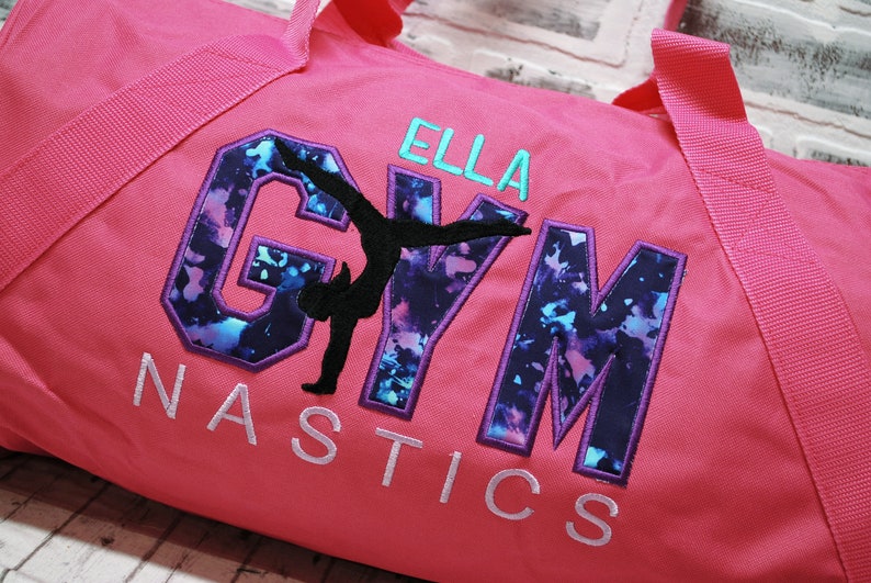 May include: Pink gym bag with a purple and blue "GYM" design with a silhouette of a gymnast in the middle. The text "ELLA GYMNASTICS" is embroidered on the bag.