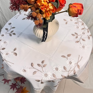 Può includere: Una tovaglia rotonda bianca con motivi floreali ricamati e un bordo decorativo. Un vaso con fiori colorati è appoggiato sul tavolo. Il vaso ha un nastro nero con la scritta "Just for you".