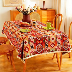 May include: A rectangular tablecloth with a geometric pattern in shades of red, orange, and beige. The design features repeating shapes and patterns, with a white fringe along the edges. The tablecloth is set on a wooden table with chairs.