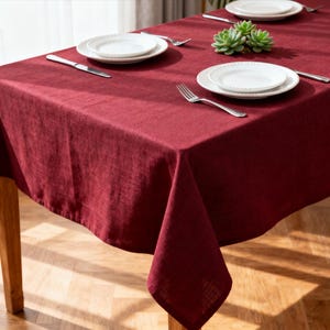 May include: A burgundy linen tablecloth covers a wooden table, set with white plates, silverware, and a small succulent. The table is set for a meal, with the tablecloth draping over the edges.