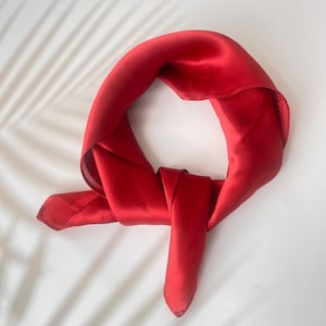 May include: A vibrant red silk scarf, knotted and arranged on a white surface. The scarf's smooth texture and rich color are highlighted by the soft lighting. The scarf is a versatile accessory.