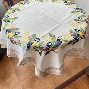 Op de afbeelding: Een ronde, crèmekleurige tafelkleed met een geborduurde rand met kleurrijke vogels, groene bladeren en rode bessen. De rand is afgewerkt met fijn kant. Het tafelkleed ligt op een ronde tafel.