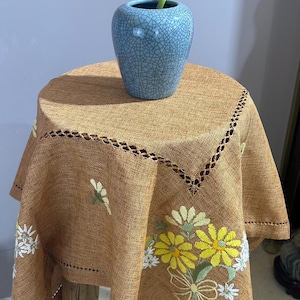 May include: A brown linen tablecloth with embroidered yellow and white flowers. The tablecloth is on a wooden table with a blue ceramic vase with a yellow flower.