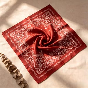 Op de afbeelding: Een vierkante, dieprode bandana met een wit paisley patroon. De stof is in een spiraal gevouwen, waardoor het ingewikkelde ontwerp wordt benadrukt. De bandana is waarschijnlijk gemaakt van zijde of een vergelijkbaar glad materiaal.