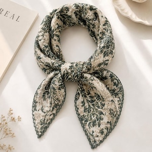 May include: A square scarf featuring a dark green and beige floral damask pattern. The fabric has a lightweight appearance and is finished with a delicate, dark green scalloped lace trim along the edges, tied in a simple knot at the centre.