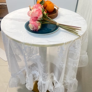 May include: A round white table with a delicate lace tablecloth. A bouquet of pink roses rests on a teal plate. A small basket of oranges sits nearby. The table has a gold trim.