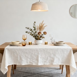 Op de afbeelding: Een eettafel gedekt met een wit linnen tafelkleed, beige borden en amberkleurig glaswerk. Een bloemstuk en kaarsen sieren de tafel. Twee beige stoelen staan aan weerszijden van de tafel. Een hanglamp hangt erboven.