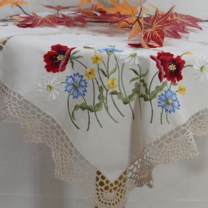 Puede incluir: Mantel crema con flores bordadas en rojo, azul, amarillo y blanco. El mantel tiene un borde de encaje de ganchillo en color beige claro. Hojas de otoño dispuestas encima del mantel.