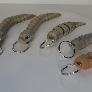 May include: Five different keychains shaped like worms with realistic details. Each keychain has a silver metal ring.