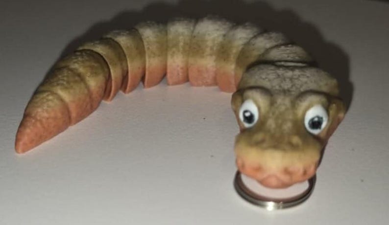 Pu&ograve; includere: Una stravagante statuetta segmentata a forma di verme, con un volto sorridente e grandi occhi espressivi. Il giocattolo &egrave; principalmente beige con accenti rosa-rossastri e sembra tenere un anello d'argento in bocca. Il design &egrave; a fumetti e giocoso.