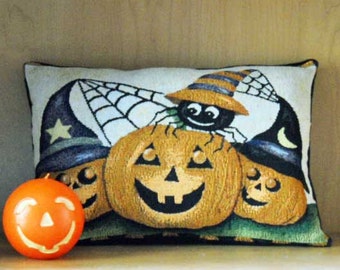 May include: A rectangular throw pillow featuring a woven Halloween scene with three jack-o'-lanterns wearing witch hats and a spider in a web, accompanied by a round orange candle with a carved jack-o'-lantern face on the left side.