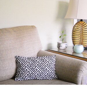 May include: A beige armchair with a black and white geometric patterned pillow. A small plant in a white pot sits on a wooden end table with a glass top. A lamp with a white shade and a wooden base shaped like a leaf sits on the table.