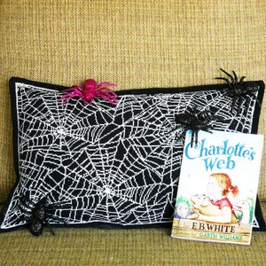 May include: A black and white spiderweb-patterned pillow with a black border, decorated with three glittery spiders. A copy of "Charlotte's Web" by E.B. White rests beside the pillow, suggesting a Halloween or spooky theme.