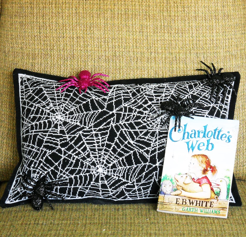 May include: A black and white spiderweb-patterned pillow with a black border, decorated with three glittery spiders. A copy of "Charlotte's Web" by E.B. White rests beside the pillow, suggesting a Halloween or spooky theme.