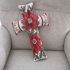 Può includere: Un cuscino decorativo a forma di croce con un motivo floreale di poinsettia. Il cuscino ha uno sfondo bianco con fiori rossi, foglie verdi e un bordo rosso. È appoggiato su una sedia imbottita beige.
