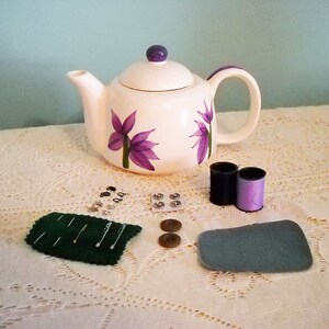 May include: A white teapot with purple flower designs, two spools of thread, a small green felt pincushion with pins, a pack of buttons, and two large buttons on a white tablecloth.