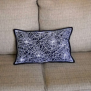 May include: A rectangular decorative pillow with a navy blue background and a white spiderweb design. The pillow has a black border and is placed on a beige couch. This Halloween-themed pillow adds a spooky touch to home decor.
