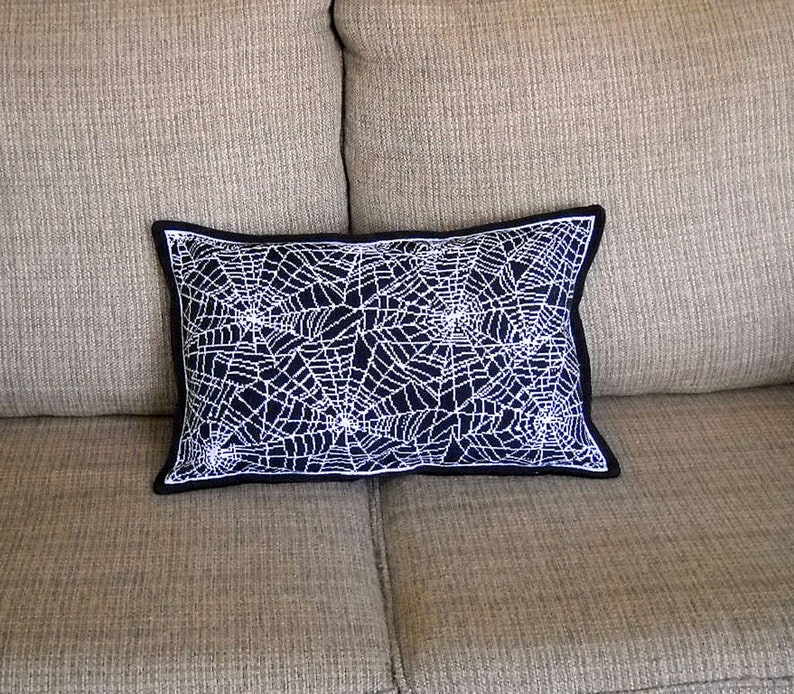May include: A rectangular decorative pillow with a navy blue background and a white spiderweb design. The pillow has a black border and is placed on a beige couch. This Halloween-themed pillow adds a spooky touch to home decor.