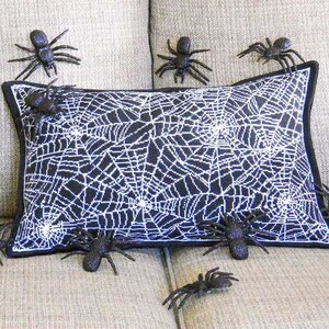 May include: A decorative Halloween pillow with a black and white spiderweb design. Several black plastic spiders are placed around the pillow, creating a spooky, festive look. The pillow has a black border.