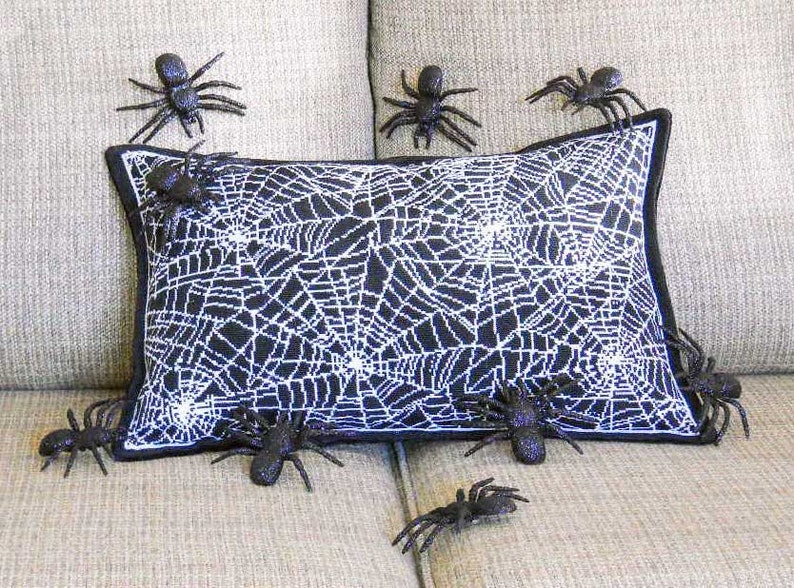 May include: A decorative Halloween pillow with a black and white spiderweb design. Several black plastic spiders are placed around the pillow, creating a spooky, festive look. The pillow has a black border.