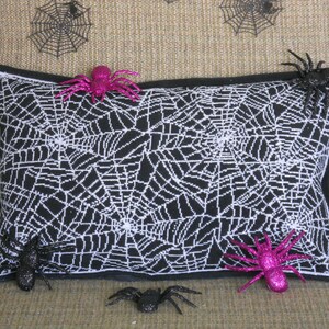 May include: A rectangular black pillow with a white spiderweb design, accented with a black border. Glittery pink and black spiders are placed on and around the pillow, creating a Halloween theme.