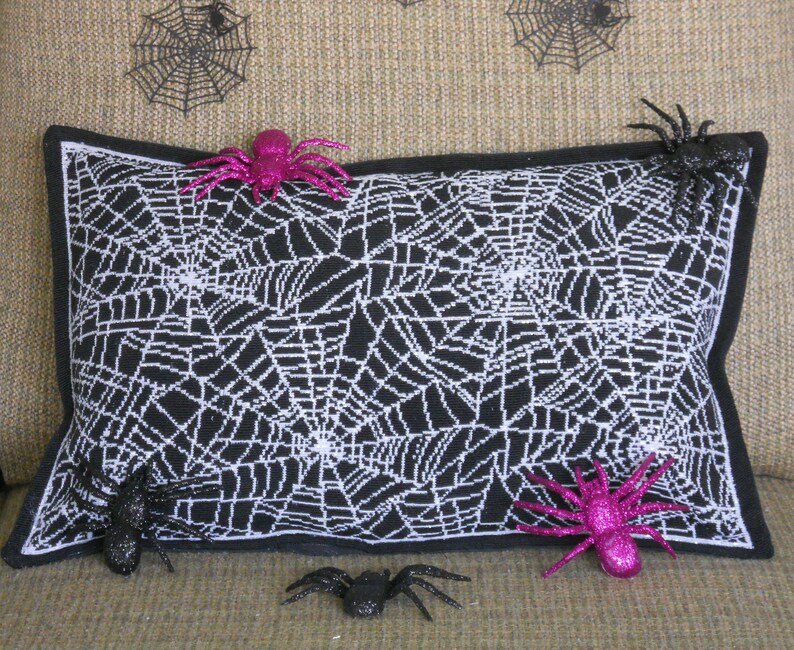 May include: A rectangular black pillow with a white spiderweb design, accented with a black border. Glittery pink and black spiders are placed on and around the pillow, creating a Halloween theme.