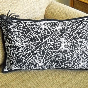 May include: Black rectangular pillow with a white spiderweb design. A black spider is attached to the upper left corner. The pillow has a black border and is placed on a beige couch. Halloween decor.