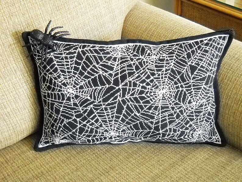May include: Black rectangular pillow with a white spiderweb design. A black spider is attached to the upper left corner. The pillow has a black border and is placed on a beige couch. Halloween decor.