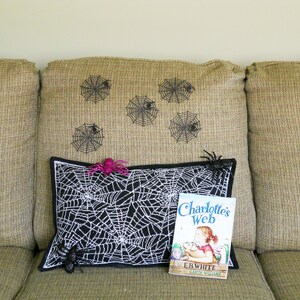 May include: A black and white spiderweb pillow with a black border, a pink spider, and a copy of "Charlotte's Web" by E.B. White. Decorative spiderwebs are on the tan couch back.