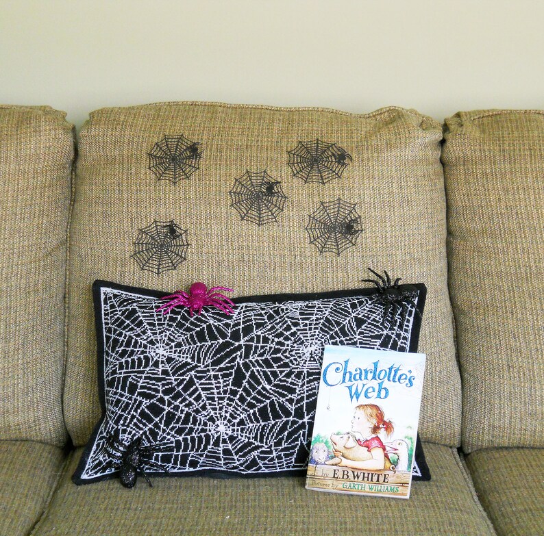 May include: A black and white spiderweb pillow with a black border, a pink spider, and a copy of "Charlotte's Web" by E.B. White. Decorative spiderwebs are on the tan couch back.
