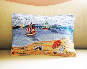 May include: Rectangular decorative pillow with a beach scene. The pillow features a blue ocean with sailboats, a sandy beach, and an orange umbrella. The pillow is made of a woven fabric.