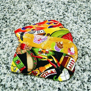 May include: A set of four colorful fabric pot holders with various designs, including a red and yellow flame, a green and yellow floral pattern, a black and white chili pepper design, and a yellow and brown cupcake design. The pot holders are made of fabric and have a quilted texture.