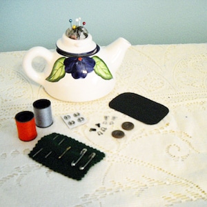 May include: White ceramic teapot with blue and green floral design, used as a pincushion with colorful pins. The teapot is on a white lace tablecloth with a black leather pad, spools of thread, safety pins, and buttons.