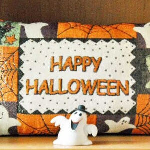Può includere: Un cuscino rettangolare a tema Halloween con un centro bianco con la scritta "HAPPY HALLOWEEN" in arancione. Il cuscino presenta motivi di ragnatele arancioni e nere, fantasmi e cappelli da strega. Una piccola statuina di fantasma è davanti.