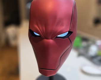 May include: A matte red full-head helmet featuring a smooth, contoured surface with black recessed panel lines and glowing white eye lenses. The helmet is mounted on a black circular display stand with a small red emblem at the base.