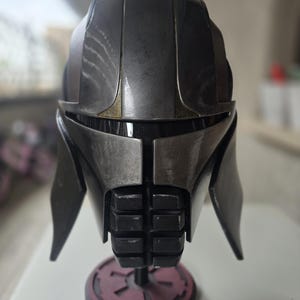 Può includere: Un elmetto dettagliato e dall'aspetto usurato nei toni del grigio e del nero, con accenti metallici. L'elmetto è esposto su un supporto nero con base rossa. Il design presenta forme angolari e una parte inferiore del viso segmentata.