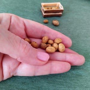 Puede incluir: Una mano sostiene patatas en miniatura de color marrón claro. Una pequeña caja de madera llena de patatas similares se encuentra en el fondo sobre una superficie verde. Las patatas son pequeñas, con una textura ligeramente rugosa.