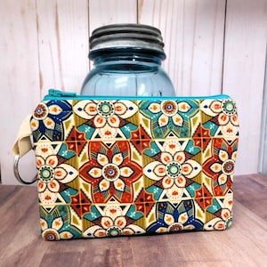 May include: A small zippered pouch with a colorful geometric pattern featuring blue, green, orange, red, and white. The pouch has a cream-colored strap with a silver ring.