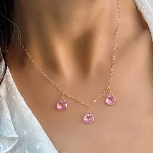 May include: A delicate gold chain necklace with three pink teardrop-shaped gemstones dangling from it.