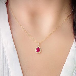 May include: A gold necklace featuring a teardrop-shaped, ruby-red pendant. The pendant is encircled by a halo of small, clear stones. The necklace is worn by a person wearing a white shirt.