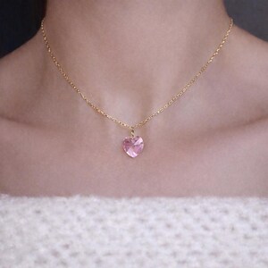 以下が含まれることがあります： ピンクのハート型のペンダントが付いた繊細なゴールドカラーのネックレス。ペンダントは、ファセット加工されたライトピンクのクリスタルです。ネックレスは人の首にかけられており、ジュエリーのデザインと色を際立たせています。