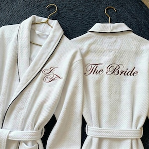 Könnte beinhalten: Zwei weiße Waffel-Bademäntel mit schwarzem Besatz und passenden Gürteln hängen an goldenen Kleiderbügeln. Auf einem Bademantel sind die Initialen "J & F" in Burgunderrot gestickt, auf dem anderen "The Bride" auf dem Rücken.