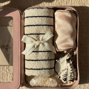 Puede incluir: Un estuche de viaje rosa con cremallera que contiene una toalla beige enrollada con rayas negras, atada con una cinta blanca. También se incluye una bolsa de satén rosa, una vela con forma de concha y una pinza para el pelo con la letra "A".