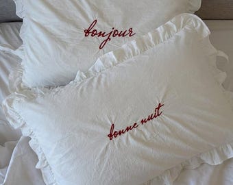 May include: Two white ruffled pillowcases with red embroidered text. One pillowcase says "bonjour" and the other says "bonne nuit". The pillowcases are on a white bedsheet, creating a cozy and inviting bedroom scene.