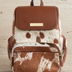 Puede incluir: Una mochila con estampado de piel de vaca en marrón y blanco y una solapa superior de cuero marrón texturizado. La solapa incluye una placa metálica rectangular de tono dorado. La bolsa tiene un bolsillo frontal con cremallera y bolsillos laterales.