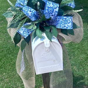 May include: A white mailbox adorned with a decorative arrangement. The arrangement includes blue and white floral patterned ribbons, green foliage, and a burlap bow. The mailbox is mounted on a wooden post and is labeled "U.S. MAIL".