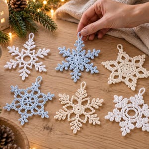 Pode incluir: Uma coleção de seis ornamentos de floco de neve feitos à mão em crochê nas cores branco, azul claro e creme. Cada floco de neve apresenta padrões intrincados semelhantes a renda e um pequeno laço no topo para pendurar sobre madeira.